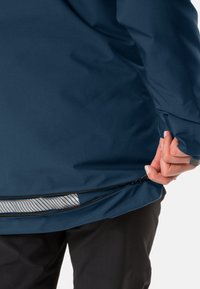 Giacca impermeabile blu scuro con zip sul fondo, dotata di una fodera interna a righe visibile attraverso l'apertura e dal taglio comodo.
