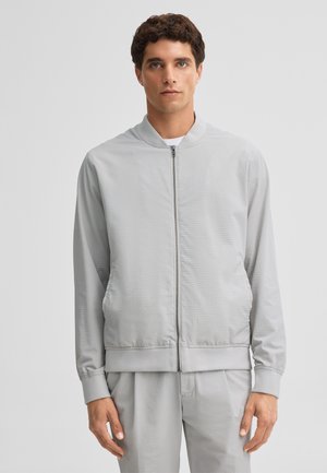 Man wearing light gray textured zip-up jacket and matching pants, standing against plain light gray background.
