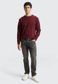 Bordeauxfarbener Strickpullover, Rundhalsausschnitt, gerippte Bündchen; dunkelgraue Jeans; weiße Sneakers; Model steht mit den Händen in den Taschen vor einem neutralen Hintergrund.