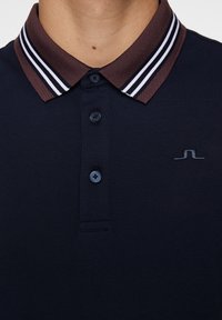 Mörkblå pikétröja med en kontrasterande krage i brunt och vitt randigt mönster, utrustad med tre knappar och en diskret logo på bröstet.