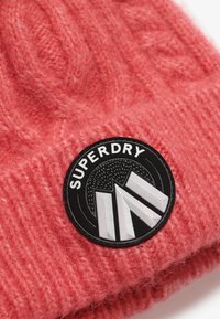 Gorro de punto rosa con un patrón de trenzas texturizadas, que presenta un parche negro circular con "SUPERDRY" y un diseño geométrico.
