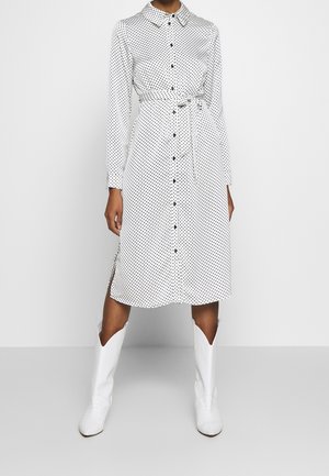 Robe blanche à manches longues avec des pois noirs, devant à boutons, col, ceinture à nouer à la taille, fentes latérales, associée à des bottes blanches de style western.