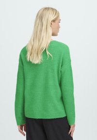 Žalias per didelis megztinis su minkštu, tekstūruotu paviršiumi. Jame yra apvalus iškirpimas, nuleistais petį ir bangelėmis apdirbti rankogaliai bei apatinė dalis.