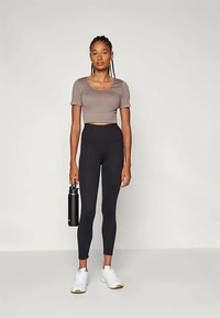 Graues Crop-Top kombiniert mit schwarzen hoch taillierten Leggings. Modell hält eine schwarze Wasserflasche. Weiße Sneakers ergänzen das Outfit.