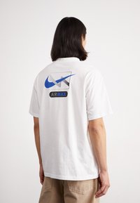 Balta trumpo rankovės marškinėliai su mėlynu Nike logotipu ir "AIR MAX" tekstu ant nugaros. Suapvalintas kaklas, laisvo stiliaus.