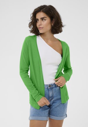 Ženska s kratkimi kodrastimi lasmi, oblečena v zeleno jopico čez belo enim ramenskim zgornjim delom in zavihane kratke jeans hlače, gleda vstran.