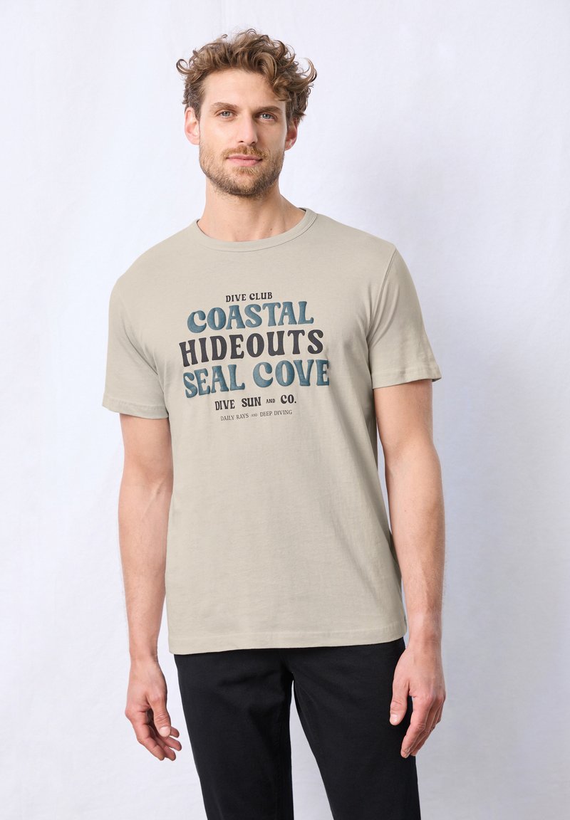 Béžové bavlněné tričko s nápisem "Potápěčský klub Pobřežní útočiště Seal Cove" vytištěným tučným námořnickým a černým písmem. Krátké rukávy a kulatý výstřih.