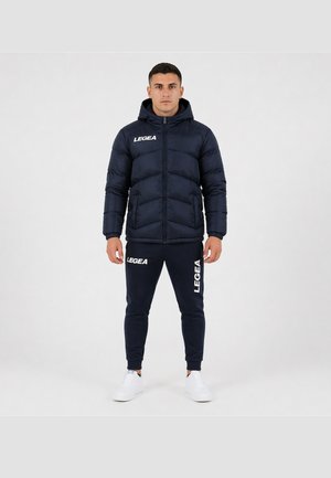 Mann steht frontal, trägt eine schwarze Steppjacke mit Kapuze und passende schwarze Jogginghose, beide mit LEGEA-Logo, und weiße Sneakers.