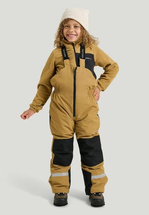 Barn iført beige og sorte isolerede snebukser med seler, en beige jakke, beige strikhue og sorte vinterstøvler, smilende med den ene hånd på hoften.
