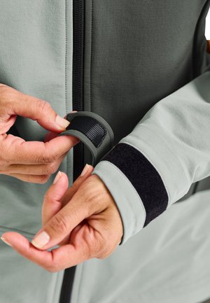 Hände, die den schwarzen Klettverschluss am Ärmel einer hellgrauen Jacke mit sichtbarem Frontreißverschluss verstellen.