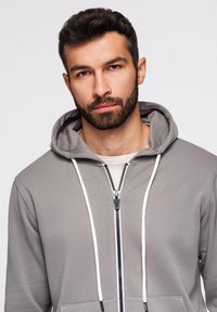 Szara bluza na zamek z białymi sznurkami, z miękkiej tkaniny, z przednimi kieszeniami i wyrazistymi detalami zamka, noszona na jasnej koszuli.