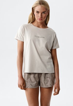 Blondynka w jasnobeżowej koszuli z napisem „LIFE IS ABOUT CHALLENGES” oraz w brązowych, wzorzystych szortach, stoi na białym tle.