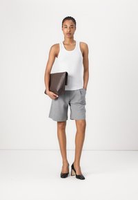 Weißes Tanktop, graue maßgeschneiderte Shorts und schwarze High Heels. Hält eine dunkelbraune Klappentasche mit geprägtem Detail. Einfache Form, minimaler Textur.