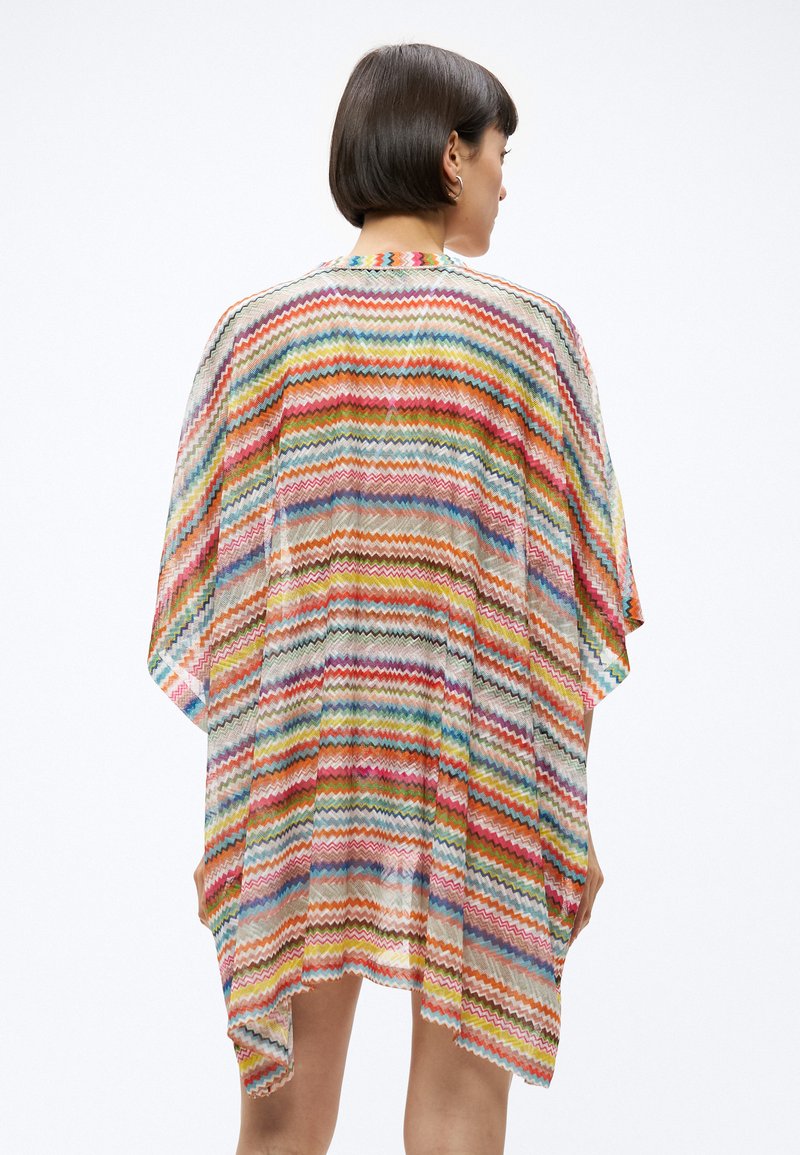 Frau mit kurzen dunklen Haaren, die ein farbenfrohes, locker sitzendes, knielanges Kimono-artiges Kleidungsstück mit Zickzackmuster trägt, von hinten auf weißem Hintergrund.