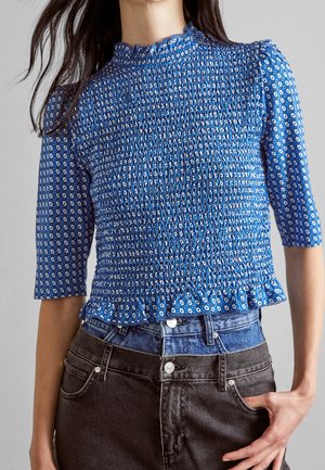 Kobieta ubrana w niebieską wzorzystą bluzkę z marszczonymi wstawkami, 3/4 rękawami i falbaną na dole, w zestawieniu z warstwowymi niebieskimi i czarnymi jeansami.