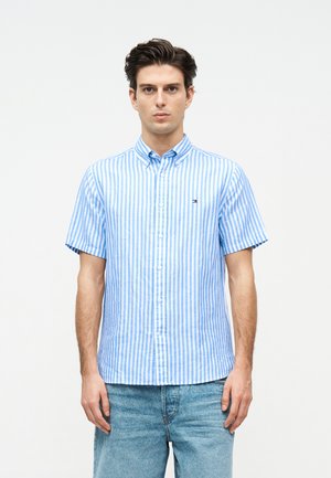 Νέος άνδρας που κοιτάζει μπροστά, φοράει μπλε και άσπρο κατακόρυφα ριγωτό κοντομάνικο πουκάμισο και ανοιχτό μπλε τζιν.