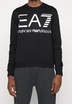 Sweat-shirt noir à manches longues avec un grand logo blanc "EA7 Emporio Armani" sur la poitrine, porté par une personne avec un pantalon foncé.