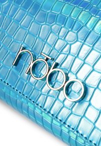 Sininen käsilaukku krokotiilikuosisella pinnalla, jossa on sileä tekstuuri, hopeinen logo ja ompeluyksityiskohdat. Ihanteellinen erilaisiin tilaisuuksiin.