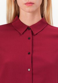Μπουρντό πουκάμισο με υφή, με μυτερό γιακά και μαύρα κουμπιά μπροστά. Δεν υπάρχουν επιπλέον μοτίβα ή στολίδια ορατά.