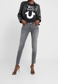 Szare wąskie dżinsy z wysokim stanem i subtelnym przecieraniem, w zestawieniu z czarną skórzaną kurtką i czarnym t-shirtem z nadrukiem oraz czarnymi sandałami na obcasie.