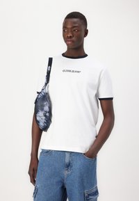 Camiseta de algodón blanca con cuello marino y logo, combinada con pantalones cortos de mezclilla azules. Llevando una bolsa de hombro de teñido anudado marino con cremallera.