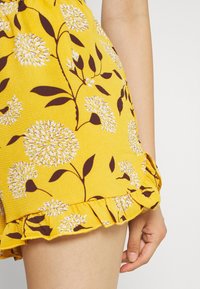Gele bloemen shorts met een gestructureerde stof, bruine bladeren en bloemenpatronen, en een gerimpelde zoom.