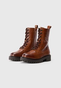 Botas de tobillo de cuero marrón con cordones, suela gruesa, punta redonda y cremallera lateral; cuentan con cordones marrones oscuros y una lengüeta.