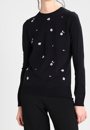 Femme portant un pull noir avec de petites fleurs blanches et violettes brodées, assorti à un pantalon noir, debout devant un fond clair.