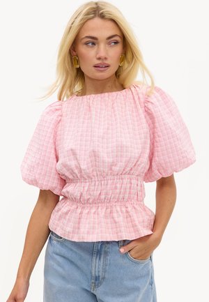 Femme blonde portant des boucles d'oreilles rectangulaires en or, un chemisier à manches bouffantes rose à carreaux vichy avec taille froncée, et un jean en denim bleu clair, main dans la poche.