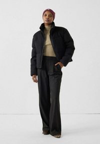 Femme portant une doudoune noire, un col roulé beige côtelé, un pantalon large noir, un bonnet bordeaux et des chaussures à motif léopard, se tenant devant un mur blanc.