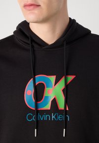 Fekete kapucnis pulóver színes logóval, amelyen kék, zöld és piros "OK" felirat, valamint kék "Calvin Klein" felirat található, fekete zsinórokkal és ezüst végekkel.