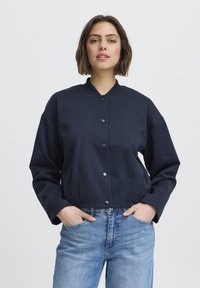 Veste cropped navy avec un col montant, des boutons pression et des manches longues ; associée à un jean en denim bleu clair. Texture douce, coupe décontractée.