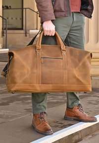 Tan leather duffel bag with dual handles and a front zip pocket. Features a smooth texture and subtle creasing; metal hardware accents.