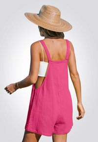 Mujer con un sombrero de paja de ala ancha, top blanco sin mangas y un mono rosa suelto sin mangas, de espaldas contra un fondo simple.