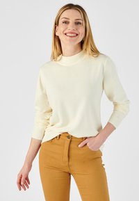 Pull crème avec un col côtelé haut et des manches longues, associé à un jean ajusté jaune moutarde, sur fond uni.