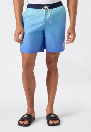 Mann trägt blaue Badehose mit Farbverlauf, navy Bund und weißem Kordelzug, kombiniert mit schwarzen Champion Slide-Sandalen.