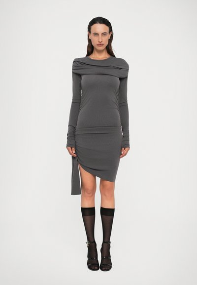 Donna che indossa un vestito grigio a maniche lunghe aderente, con orlo asimmetrico e dettaglio a incrocio, abbinato a calze trasparenti al ginocchio e tacchi neri.