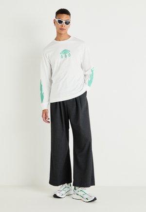 T-shirt blanc à manches longues avec un graphique d'œil vert et un design dégoulinant ; associé à un pantalon sombre ample et des baskets blanches avec des accents verts.