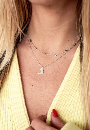 Collane a strati d'argento con ciondoli a forma di luna crescente e cuore, caratterizzate da un design di catena delicata, indossate sopra un maglione giallo strutturato.