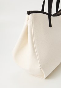 Borsa tote in tela di colore crema con bordi neri. Presenta una forma strutturata, due manici in tessuto e una superficie texturizzata. Nessun hardware visibile.