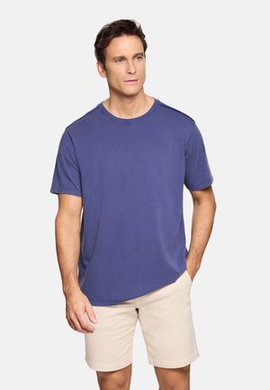 Hombre de pie con la mano en el bolsillo, vistiendo una camiseta azul lisa de manga corta y pantalones cortos beige sobre un fondo blanco.