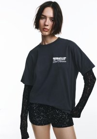 Črna bombažna majica s tiskanjem besedila, z dolgimi čipkastimi rokavi. Kombinirana s črnimi kratkimi hlačami z bleščicami, ki poudarjajo kontrastne teksture.