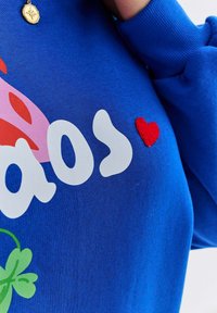 Modrá mikina s výrazným bielym textom, ružovou grafikou a malým červeným plsteným srdcom. Mäkká látka s voľným strihom.