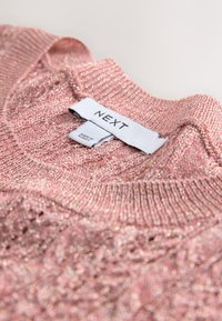 Lyserød strikket sweater med tekstureret stof, med ribbet halsudskæring og en mærkat med "NEXT"-mærke. Diskret glimmer over det hele.
