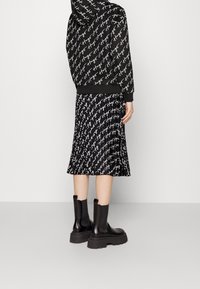 Persona vistiendo botas negras hasta la mitad de la pantorrilla, una falda plisada negra y una sudadera negra con un patrón de texto en blanco repetido.