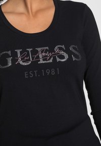 Zwarte lange mouwen shirt met een zilveren strassontwerp met "GUESS," "Los Angeles," en "EST. 1981" in een textuurdruk.