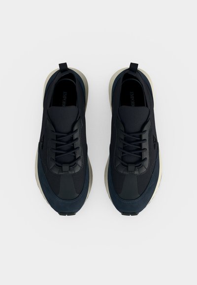 Paire de baskets de sport noires à lacets avec semelles épaisses couleur crème vues de dessus sur un fond uni.