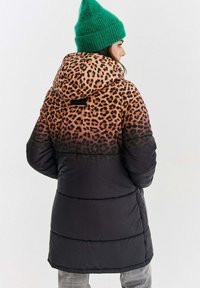 Žena stojaca bokom, oblečená v čiernej a leopardovej vzorovanej puffernovej bunde s kapucňou a jasne zelenou pletenou čiapkou.