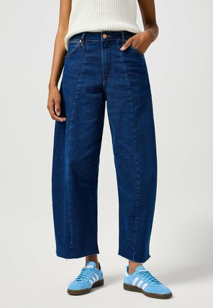 Jeans a gamba larga blu scuro con vita alta, orlo grezzo e tasche frontali, abbinati a sneakers blu chiaro con strisce bianche.