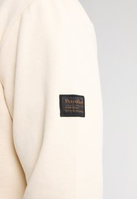 Suéter de tela beige con un acabado texturizado, que presenta una etiqueta negra en la manga con letras y detalles en dorado.